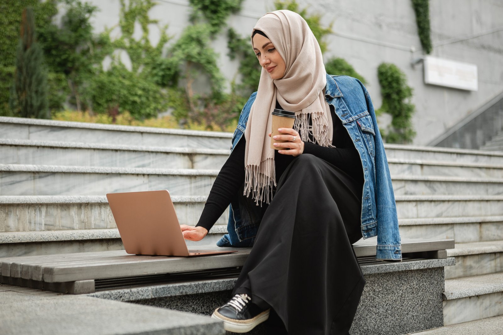 modern stylish muslim woman in hijab, denim jacket and black abaya sitting in city street working on laptop, freelancer online remote work, drinking coffee